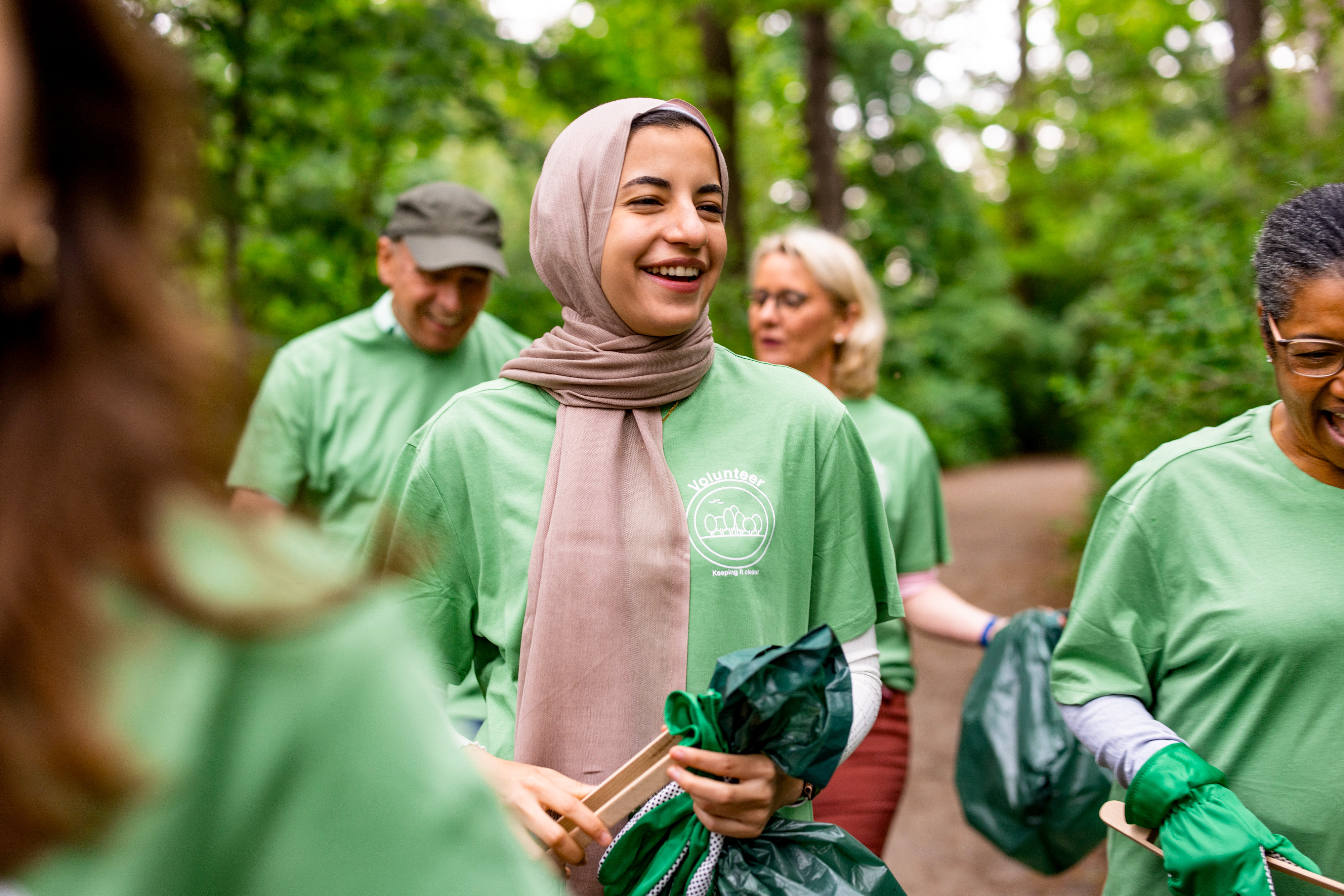 A group of volunteers wearing green shirts and holding trash bags are cleaning up a forested area.